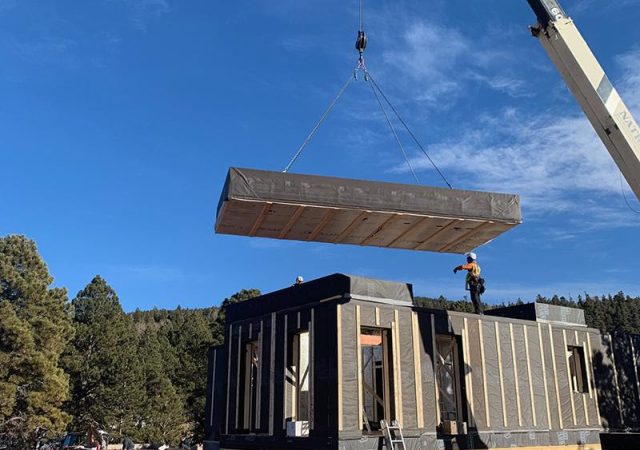 A very tall crane lowers a portion of the roof onto a prefabricated metal building.
